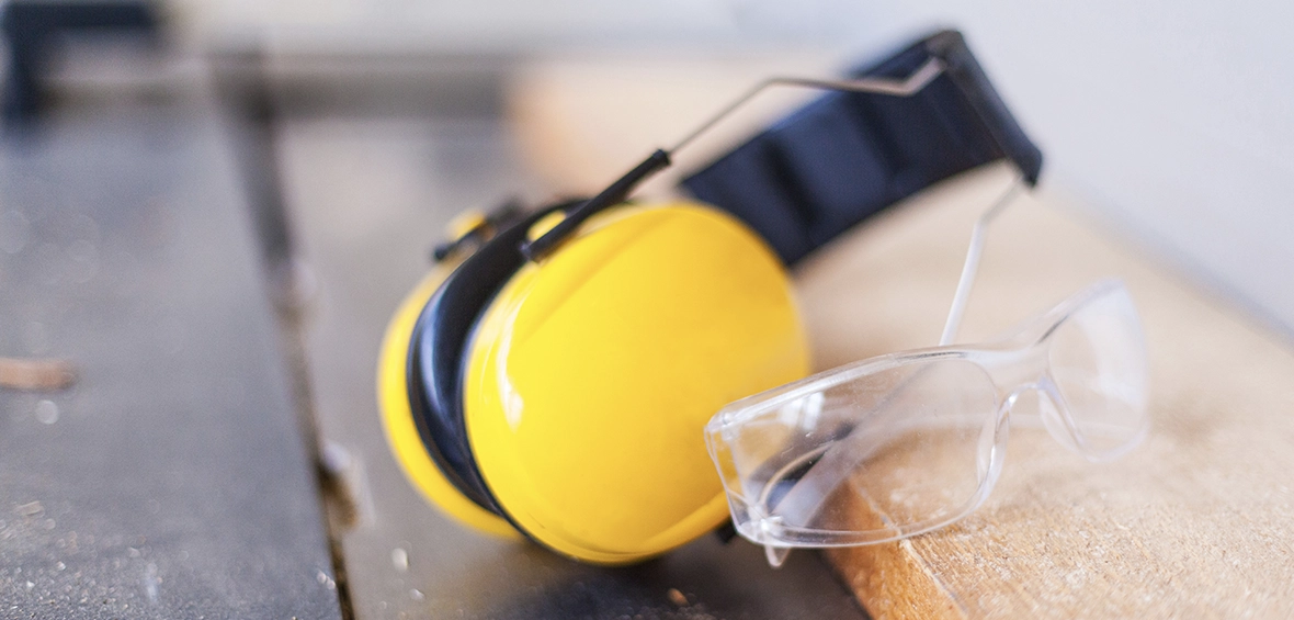 Protective ear muffs and clear safety glasses lean against a plank of wood. 