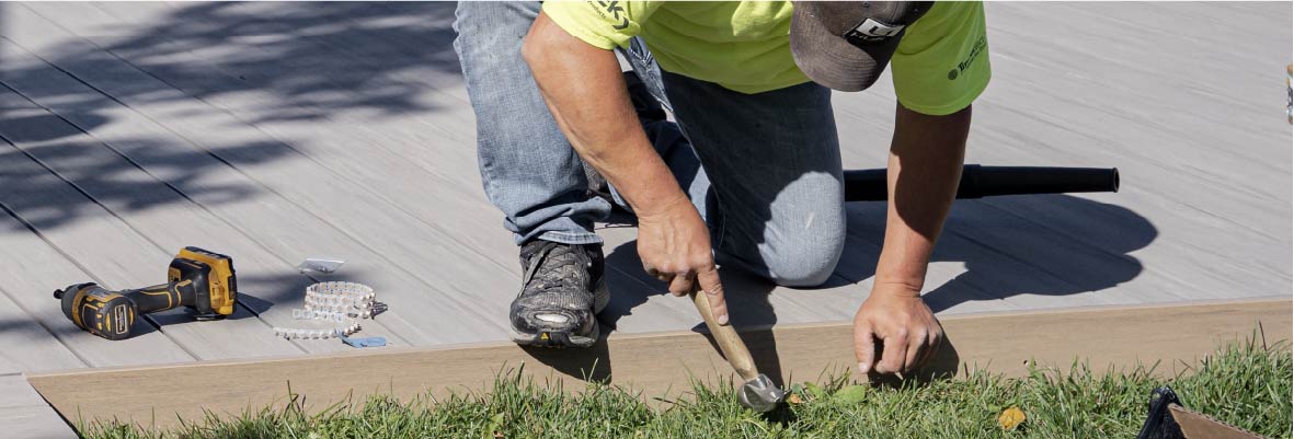 A contractor leans over the edge of a deck build to drive nails into the rim board with a hammer.