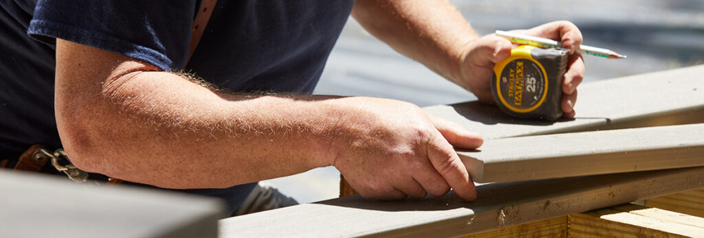 A close up photo shows a contractor adjusting a deck board with a measuring tape and pencil in hand.