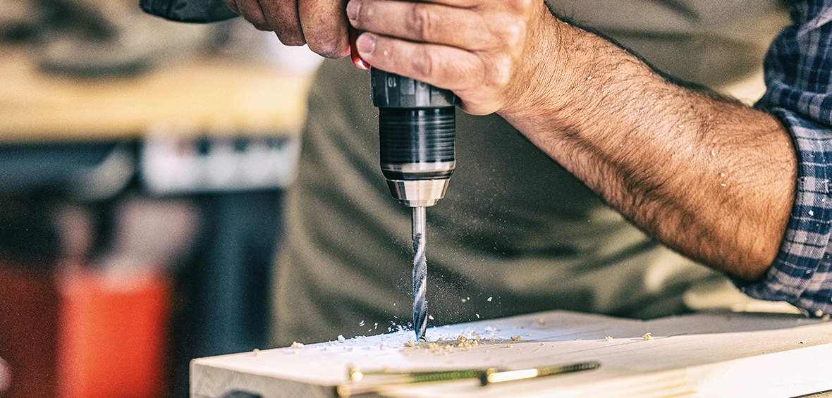 Hands holding a power drill as a large drill bit is driven through a piece of lumber. 