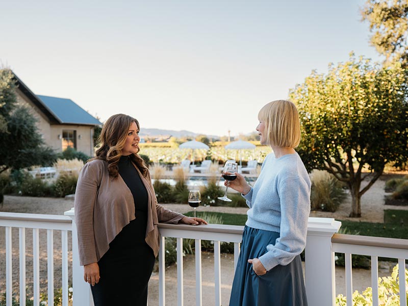 Erin stands opposite of one of her friends. The two women look engaged in the conversation and each have a glass of red wine.