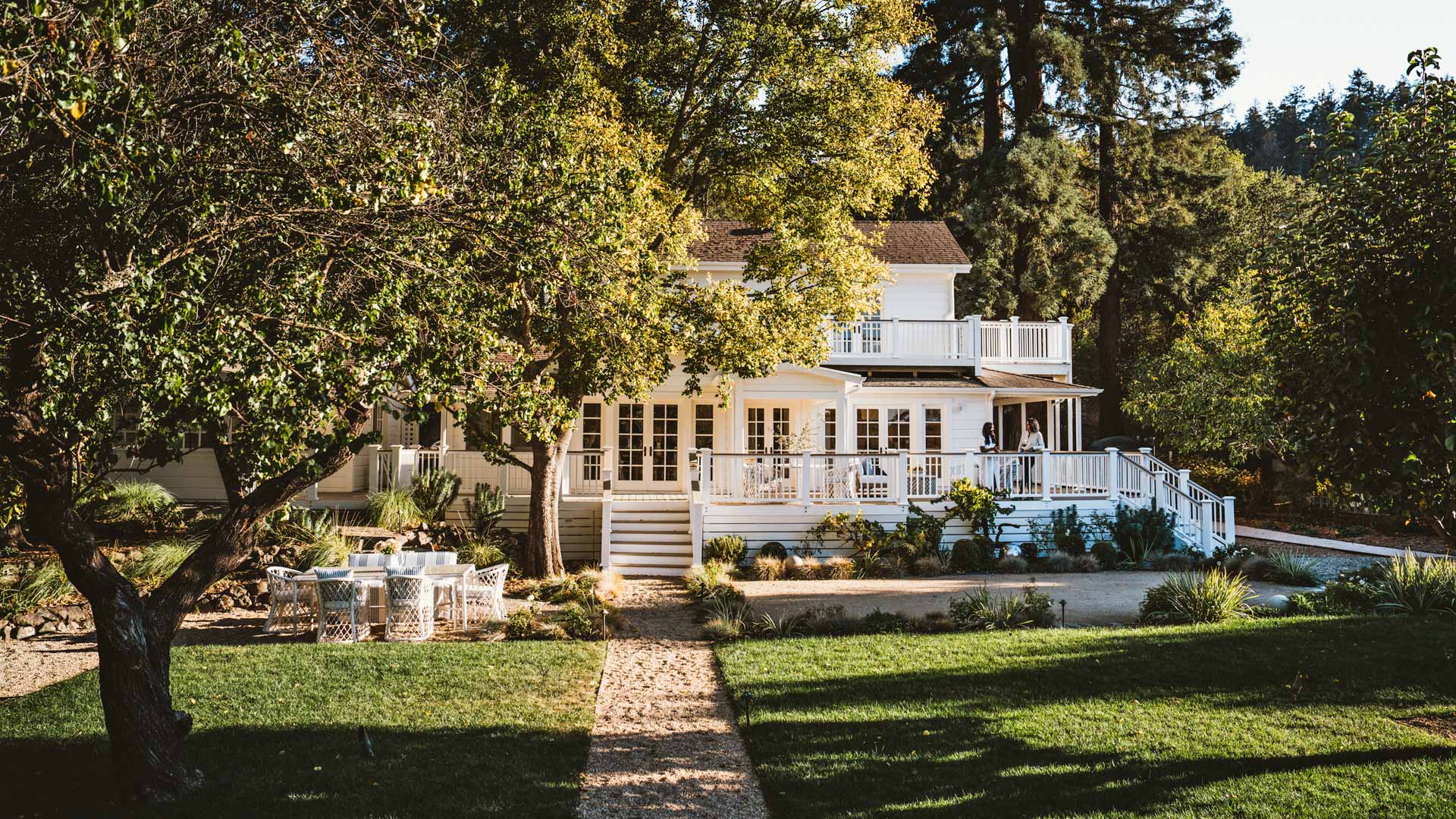 Large, white house with wrap-around porches and decks on the ground and second floor. A large deciduous tree eclipses the house in the foreground.